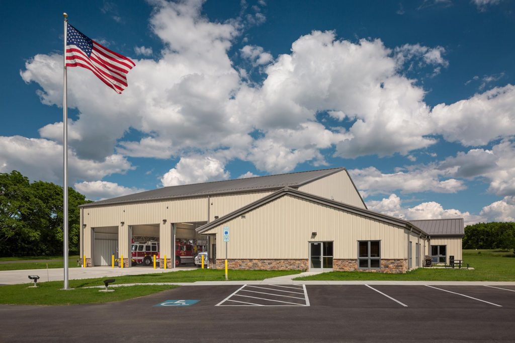 Beige metal building firehouse with 3 bay doors that are open, darker brown accents and a stone building base.